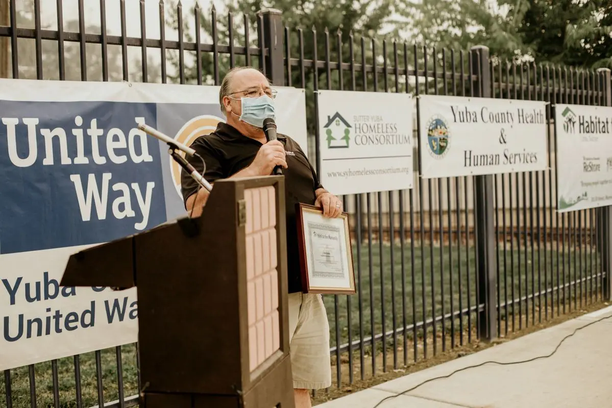 A man wearing a mask speaks at a podium in front of a fence.