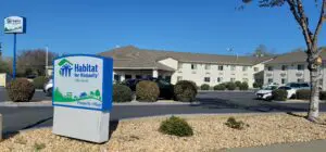 A habitat for humanity sign at the entrance of a parking lot with a multi-story building and cars parked under a clear blue sky.