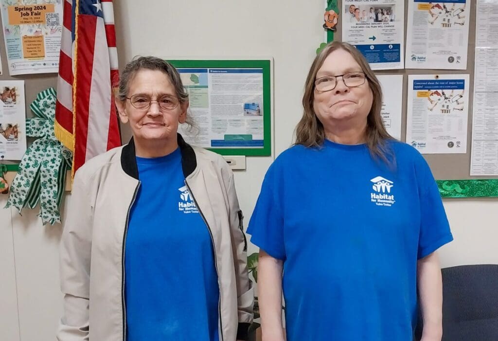 Two women in habitat for humanity t-shirts standing in an office with a bulletin board and a flag in the background.