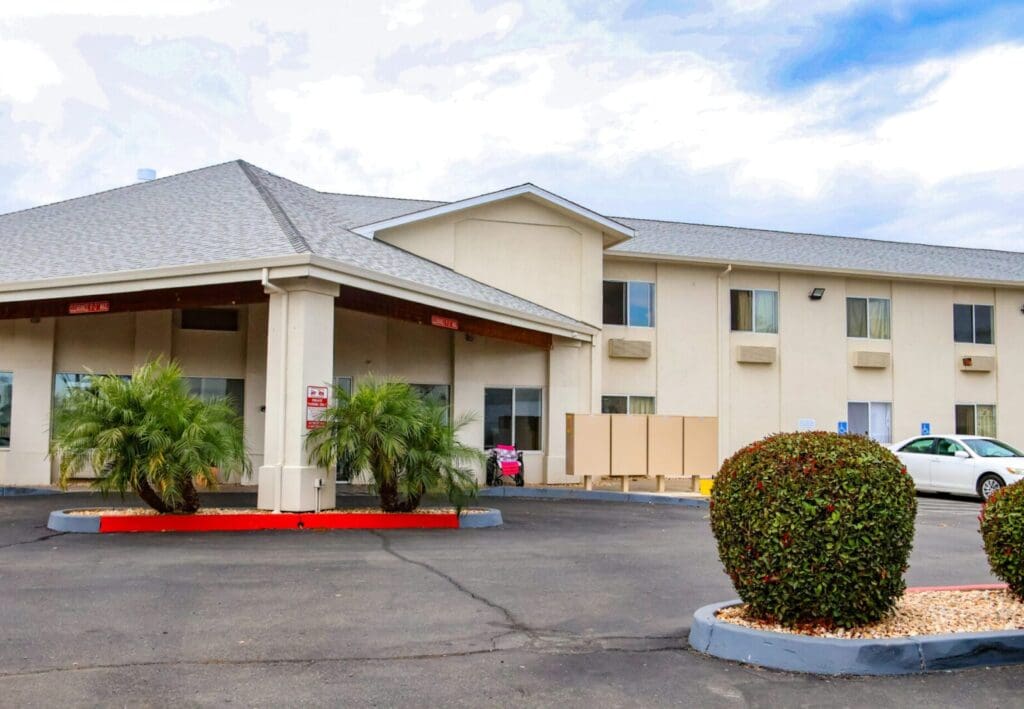 A two-story motel with a beige exterior and red trim around the windows and roof, featuring a parking lot with a few cars and trimmed bushes.