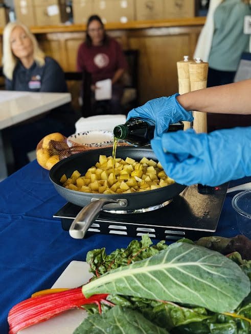 A person in blue gloves pours oil into a pan of diced potatoes at a cooking demonstration, with onlookers in the background.