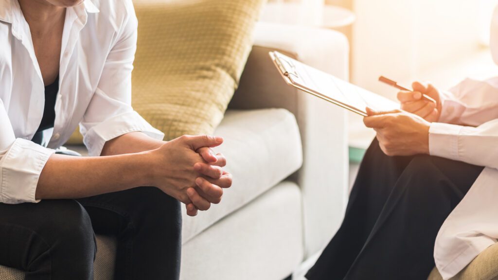 A woman with a clipboard sits with another woman with hands folded together